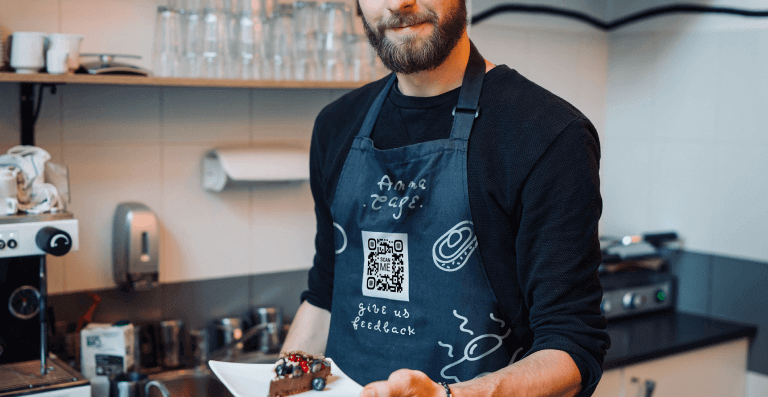A man with a cake in his hand wears an apron with a Feedback QR Code at Anna Cafe.